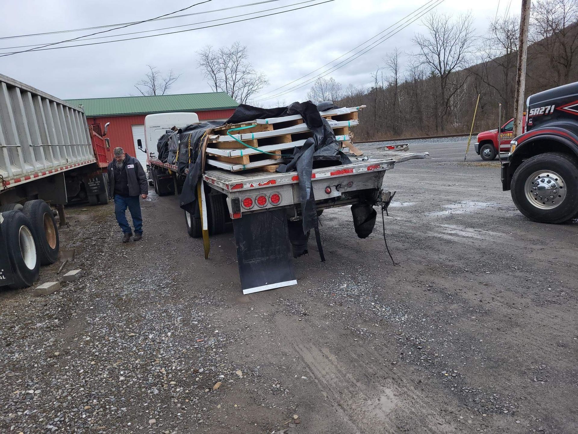 Tractor trailer loaded with lumber in a gravel yard, with people standing nearby and a red truck at right.