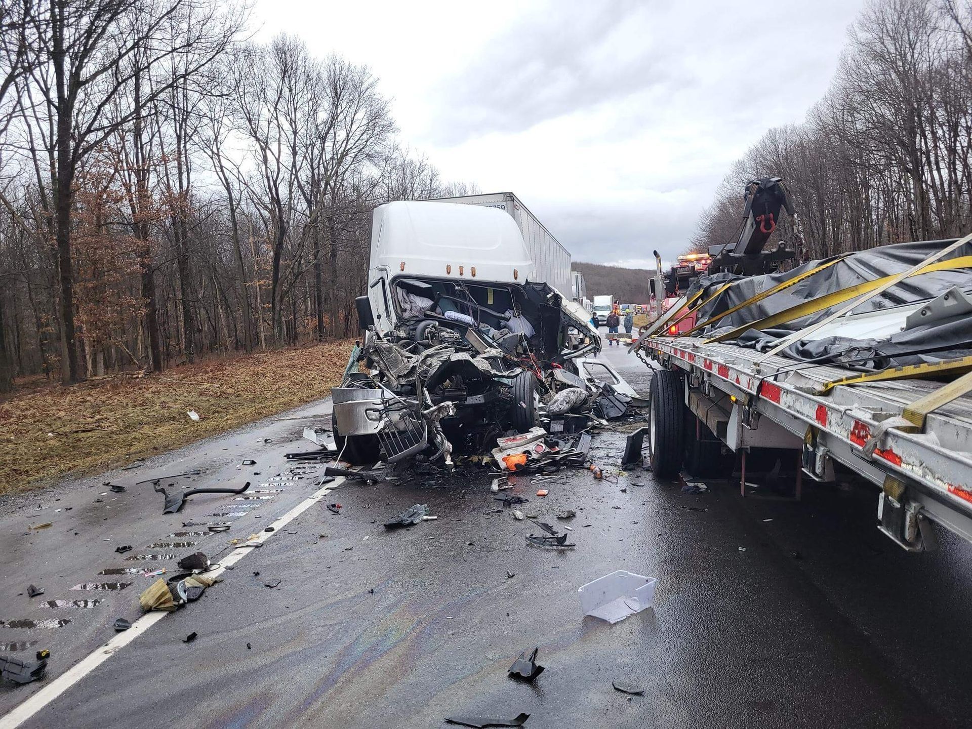 Severe highway crash with a mangled white truck and a tow truck on a wet road, debris scattered nearby