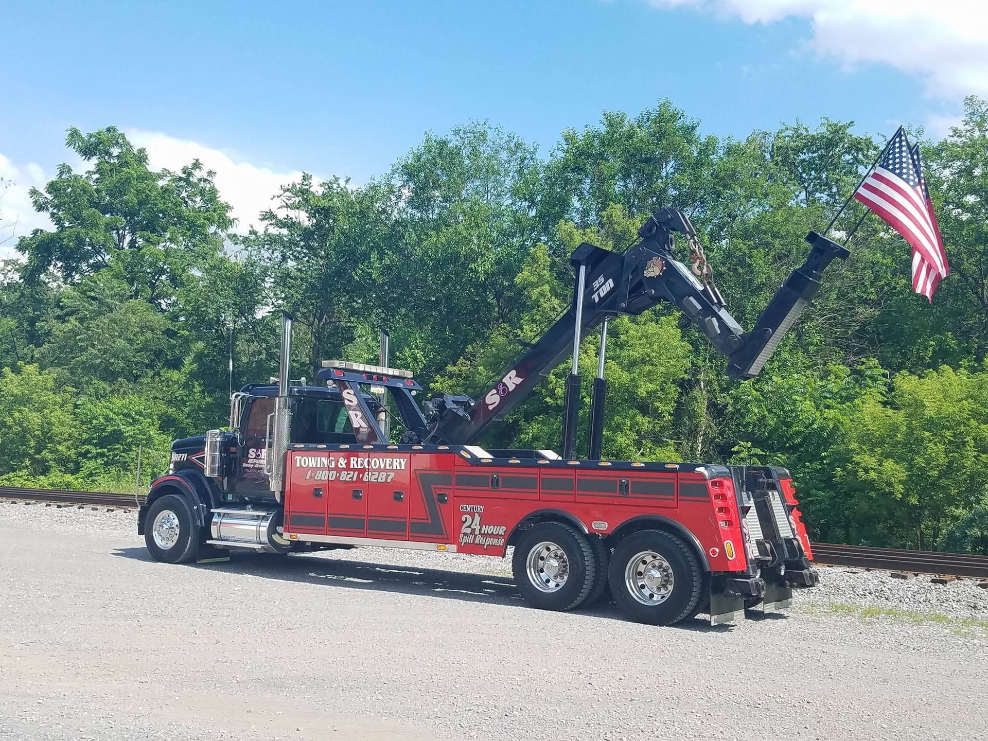 Red tow truck with raised boom parked on a roadside beside trees and an American flag.