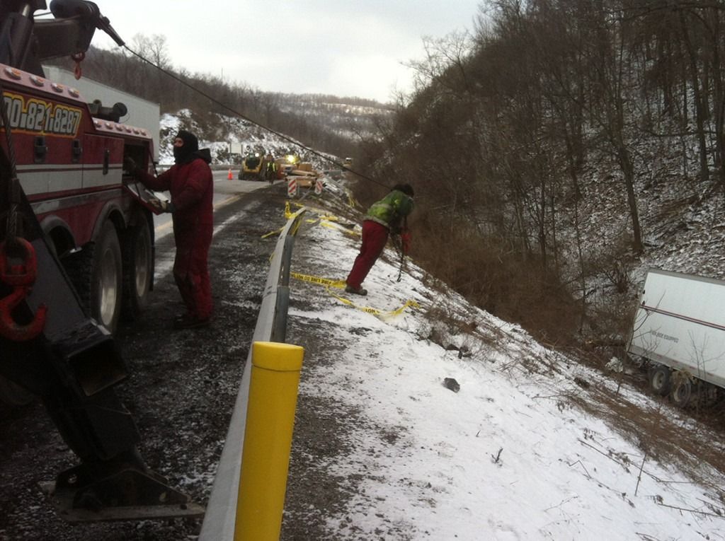Tow truck crew clearing a snowy roadside ditch beside a wooded embankment