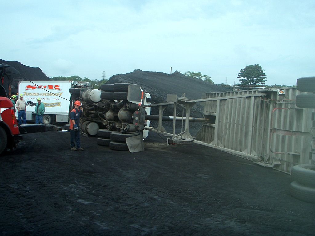 People working near a truck carrying rocks on a dark roadside, with a fence and hills in the background
