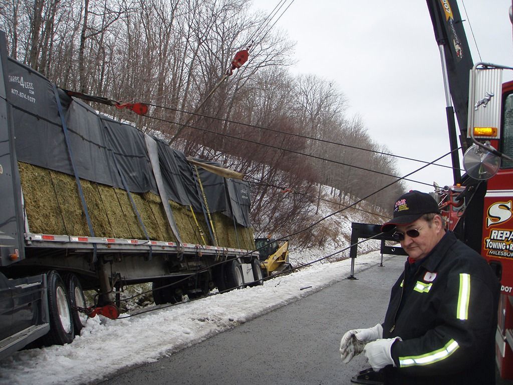 Tow truck and overturned trailer on snowy roadside, with a worker in reflective gear nearby