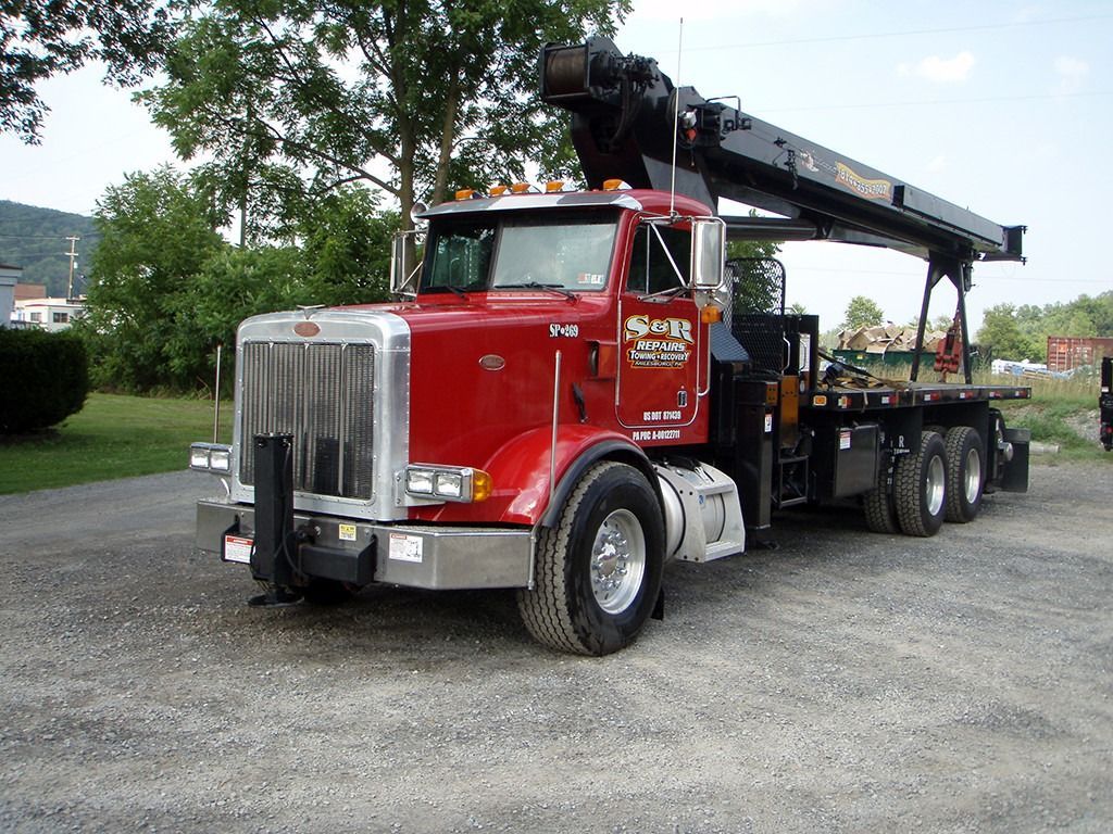 Red tow truck with crane arm parked on a road near a grassy area and trees.
