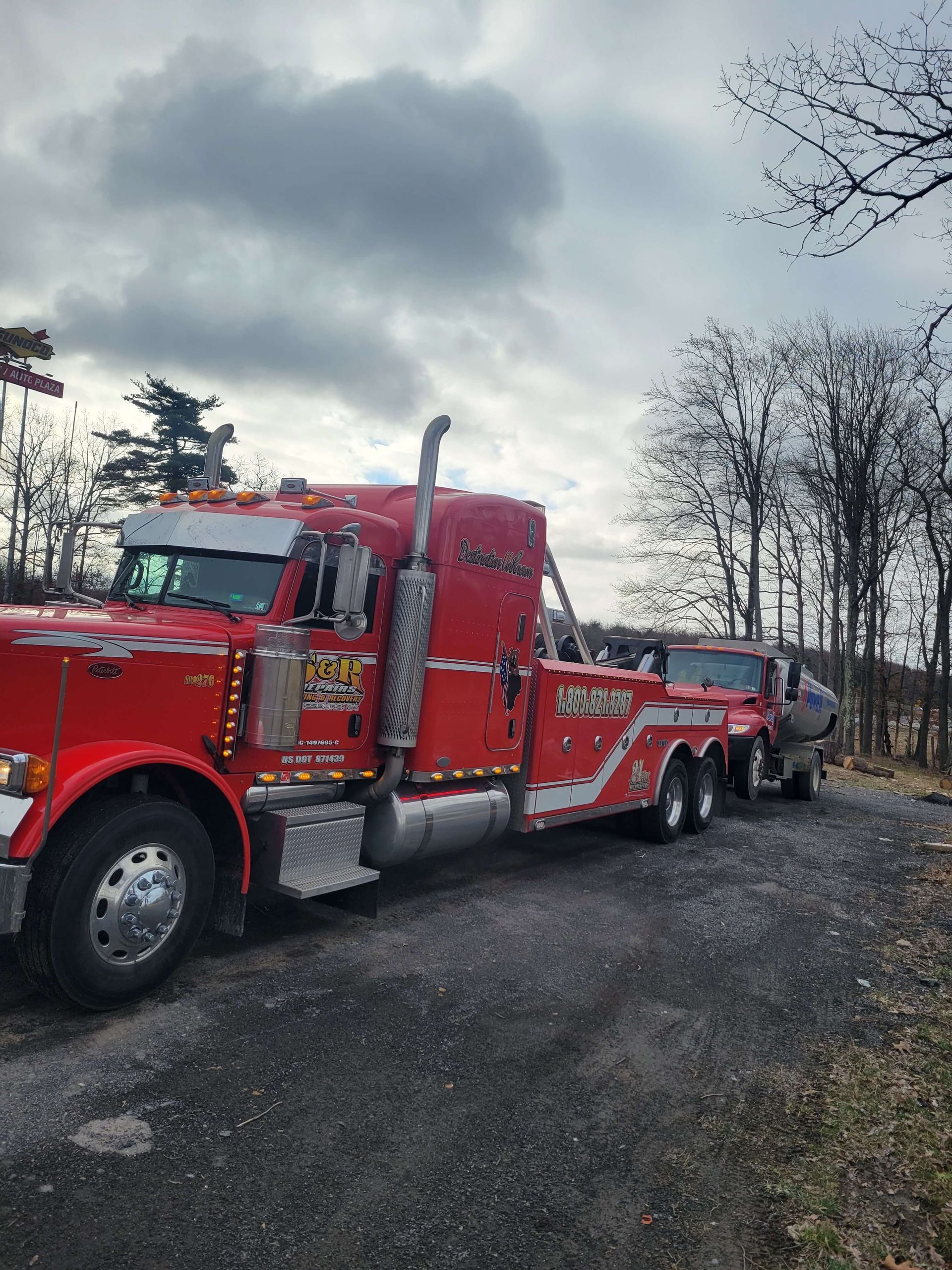 Red semi-truck with a long trailer parked on a gravel road under a cloudy sky.