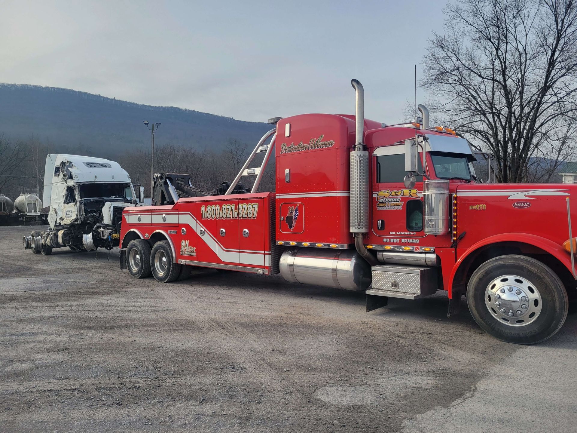 Red tow truck hauling a white semi-truck cab in a gravel lot with hills in the background.