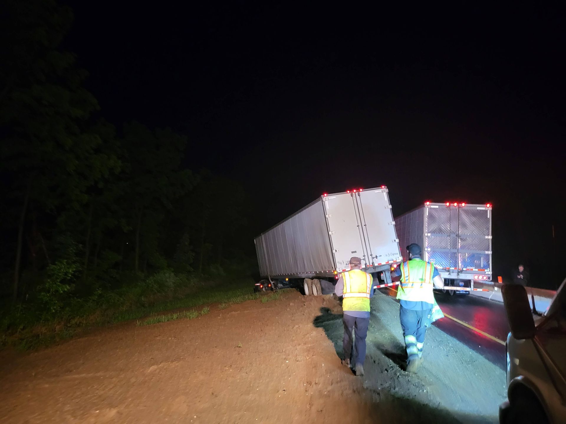 Two workers in reflective vests inspect a jackknifed semi-trailer on a dark dirt road at night.