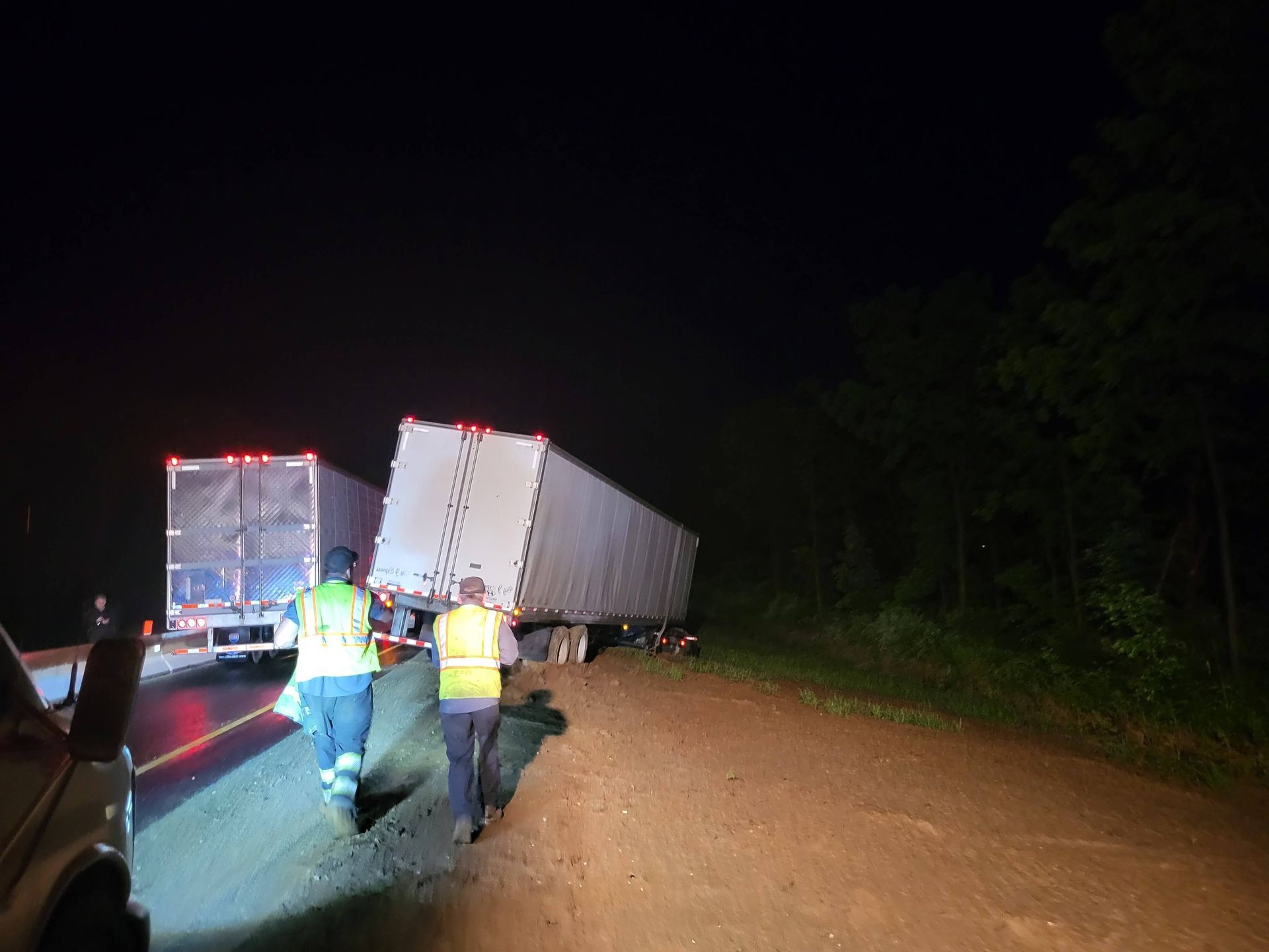 Two workers in reflective vests inspect a tipped semi-trailer on a dark dirt road at night.