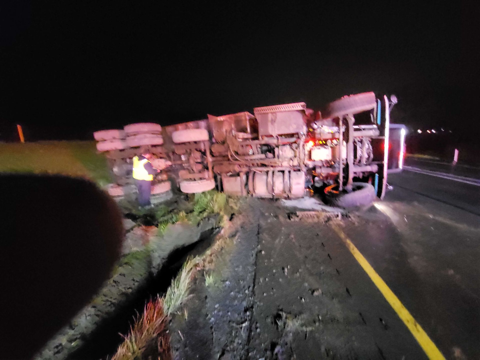 Overturned truck blocking a dark road at night, with debris scattered and emergency lights flashing nearby