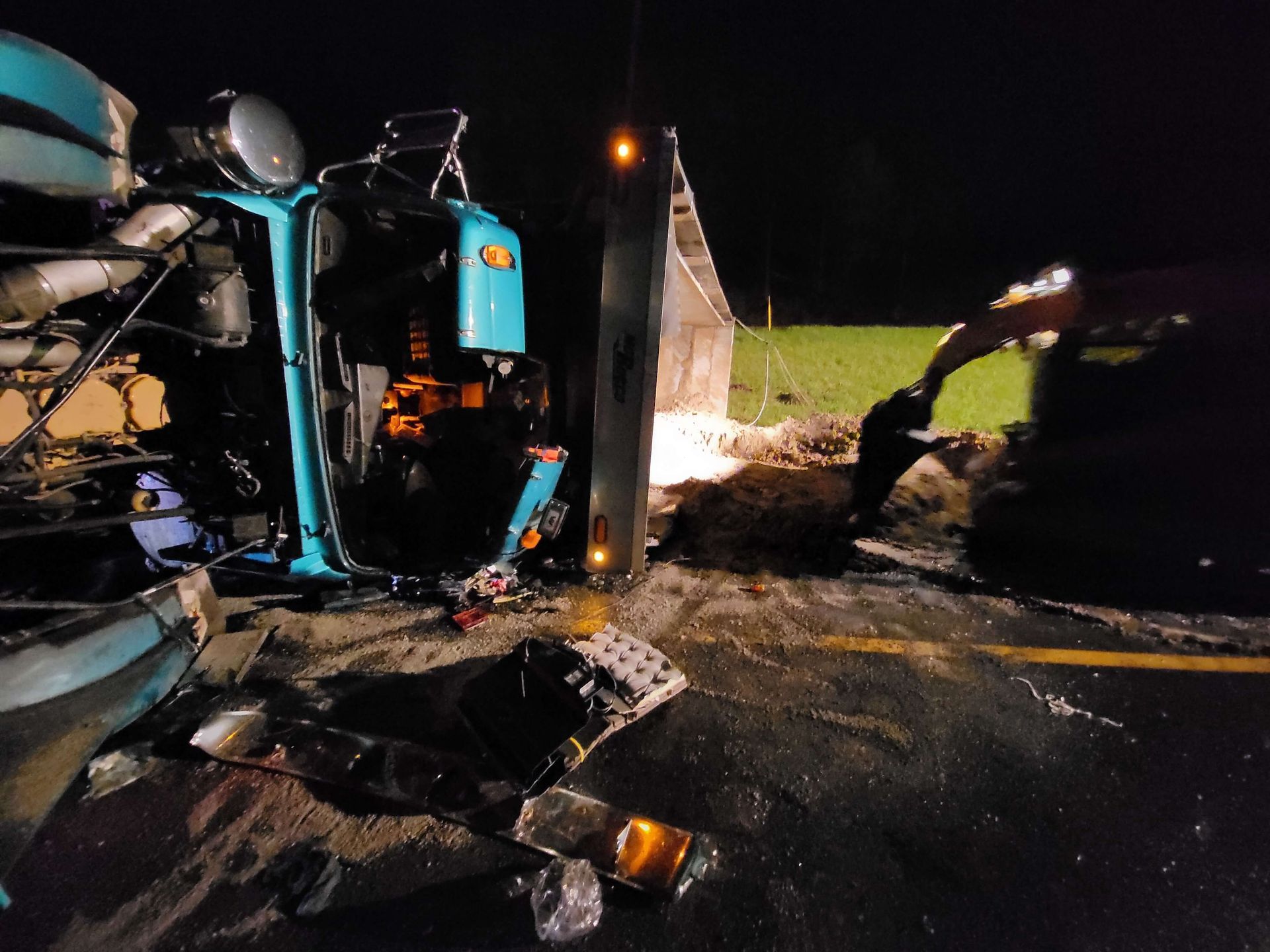 Overturned turquoise vehicle at night, with debris and flames on a wet roadside.