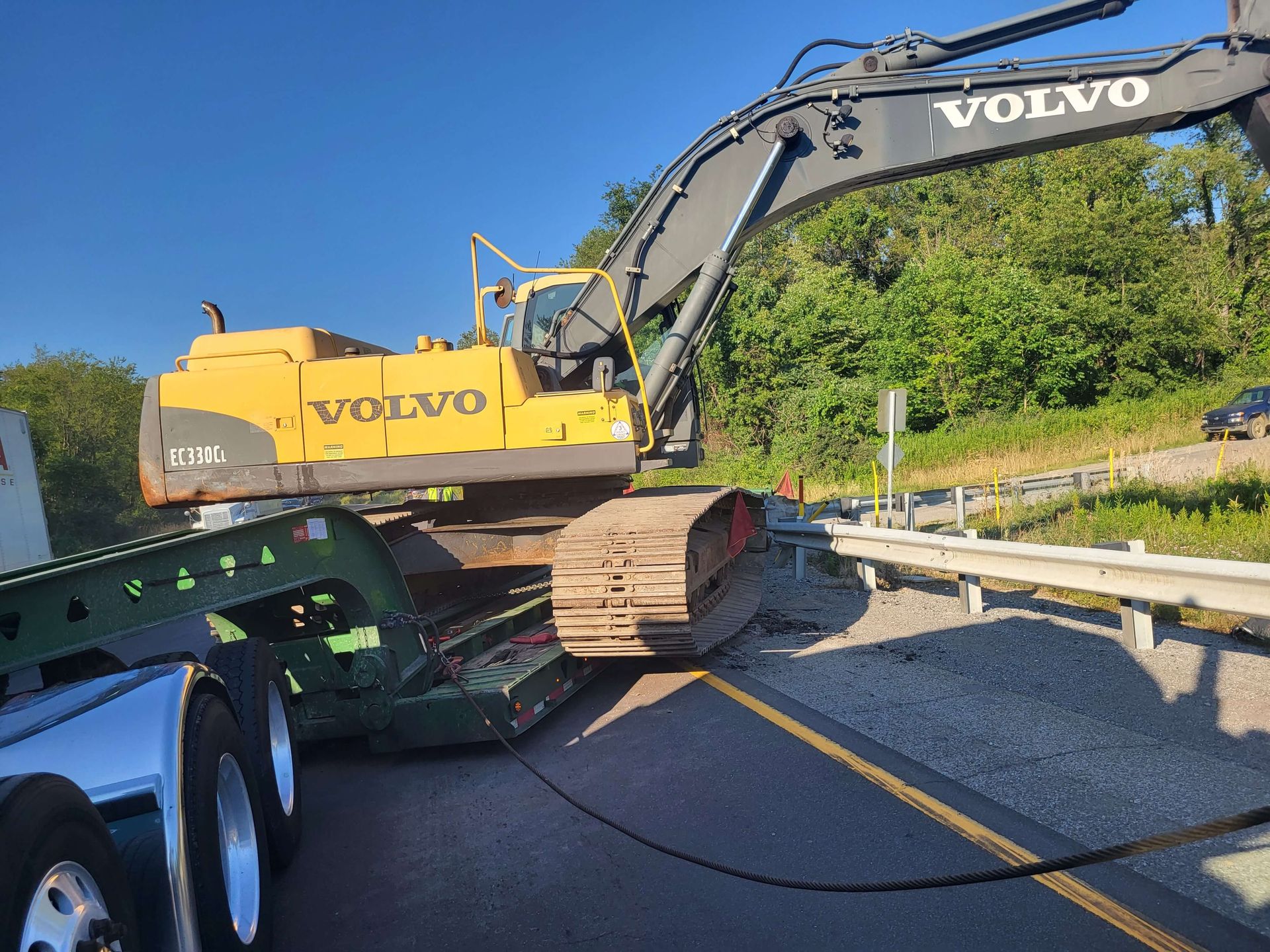 Yellow Volvo excavator on a trailer beside a road with a guardrail and trees nearby