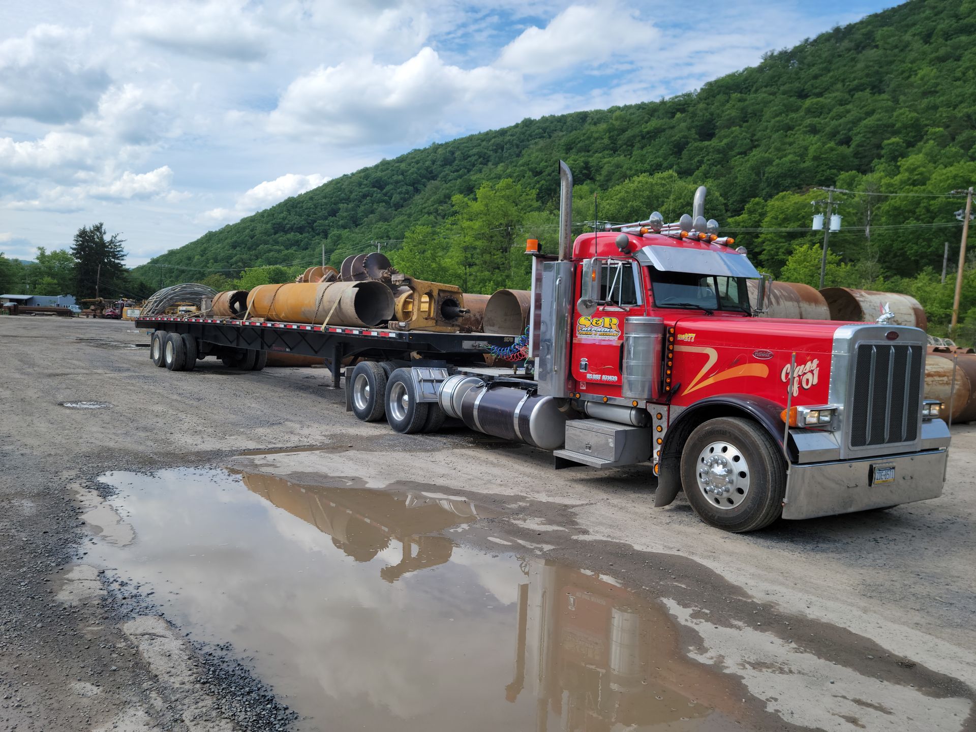 Red semi-truck hauling industrial equipment on a flatbed, parked on a muddy roadside with hills in the background