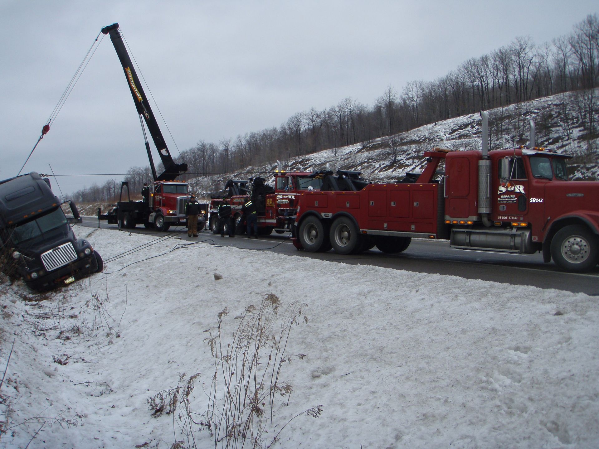 Tow trucks pulling a black car from a snowy roadside on a winter hill