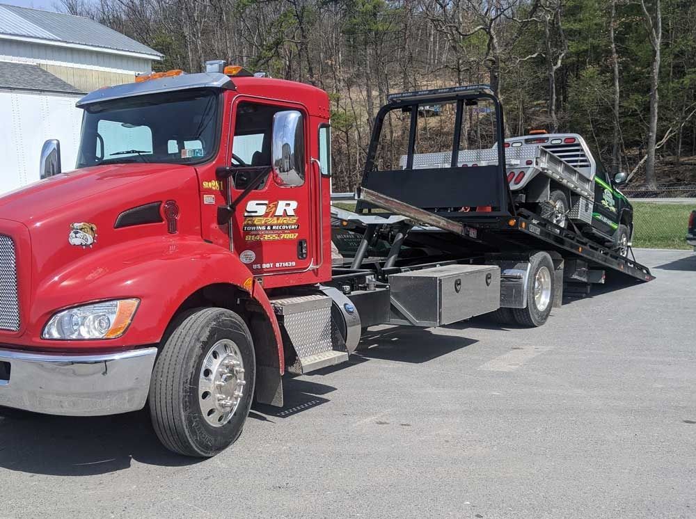Red tow truck carrying a loaded car trailer on a paved road in daylight