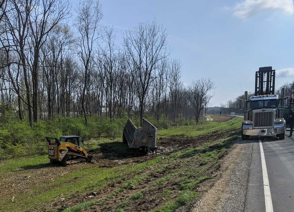 Roadside construction with a bulldozer, excavator, and truck beside a wooded embankment.