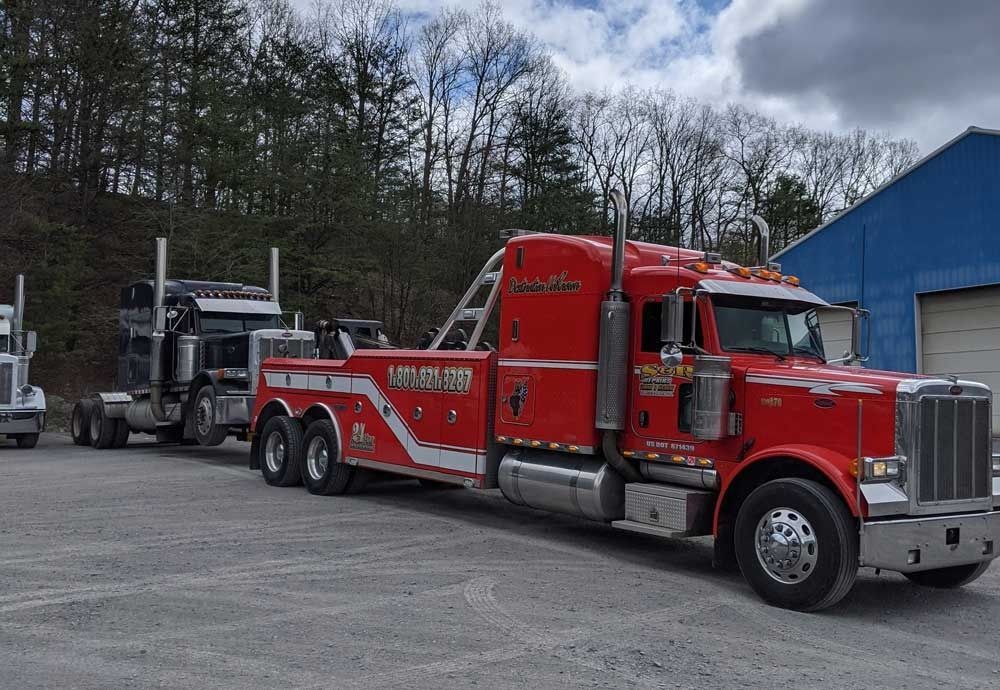 Red tow truck hauling a car trailer in a gravel lot beside a blue building