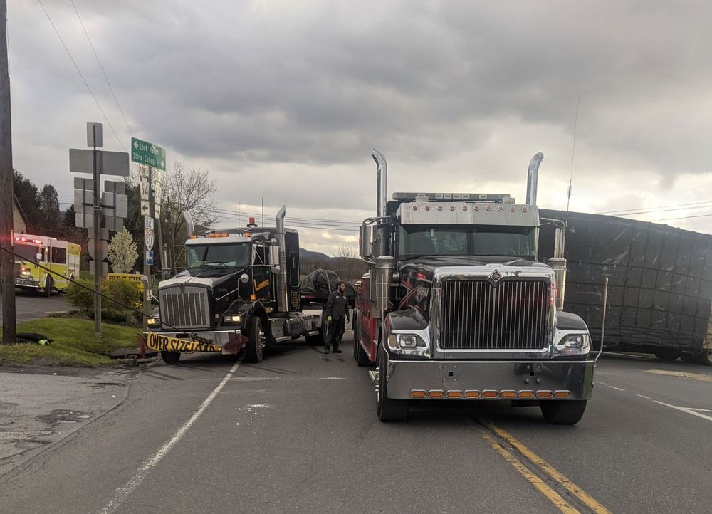 Two semi trucks blocking a wet road under cloudy skies, with one angled across the street and a damaged front bumper.