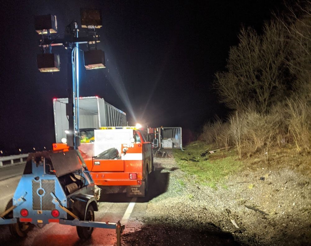 Roadside work truck with raised lights beside a gravel shoulder at night, near brush and trees.
