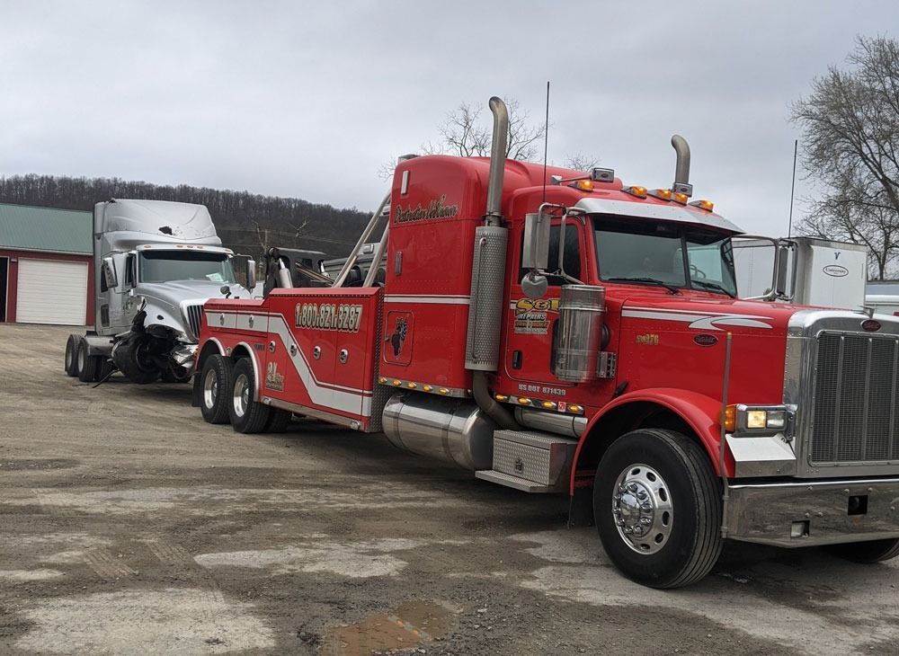Red tow truck with a white semi-truck behind it in a gravel lot on a cloudy day