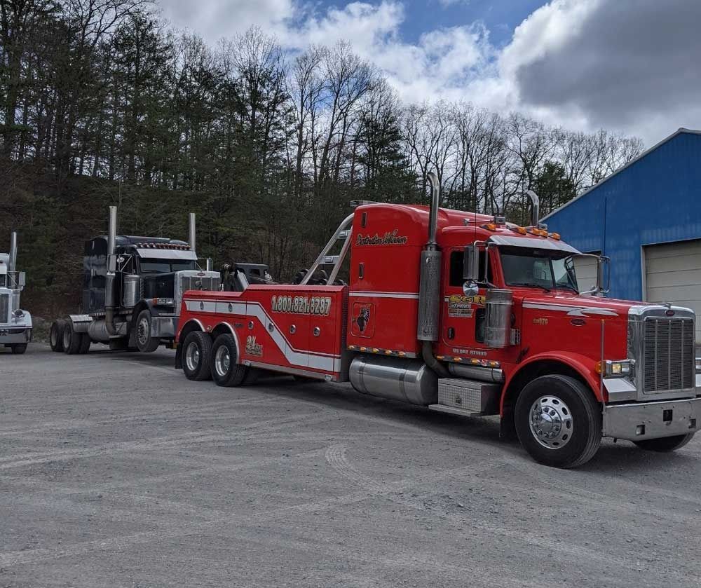 Red semi truck parked on a gravel lot beside a blue building and flatbed trailer under cloudy skies.