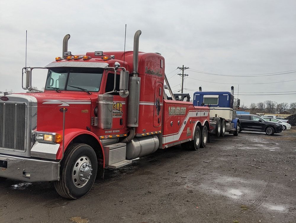 Red semi-truck towing a blue truck on a muddy lot under cloudy skies