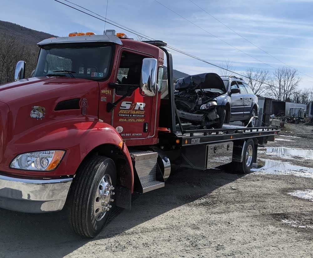 Red tow truck carrying a damaged black car on a flatbed in a snowy lot
