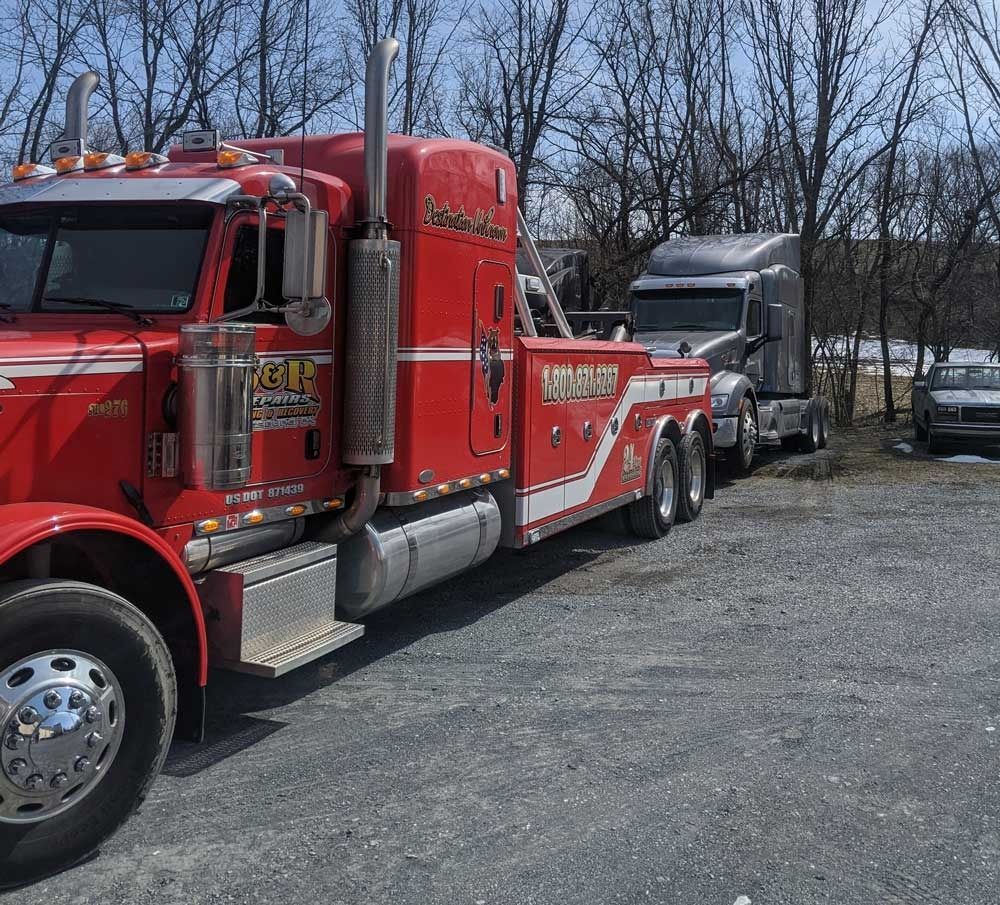 Red semi-truck towing a gray truck on a gravel lot beside leafless trees