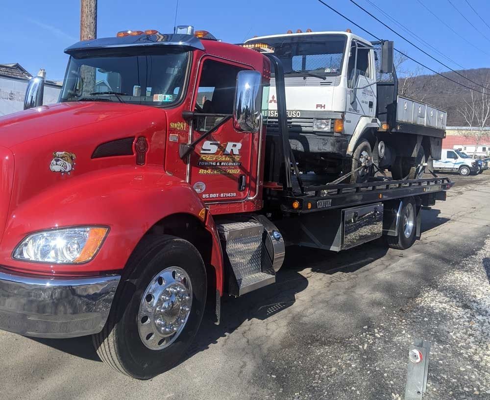 Red tow truck carrying a white dump truck on its flatbed in a sunny outdoor lot