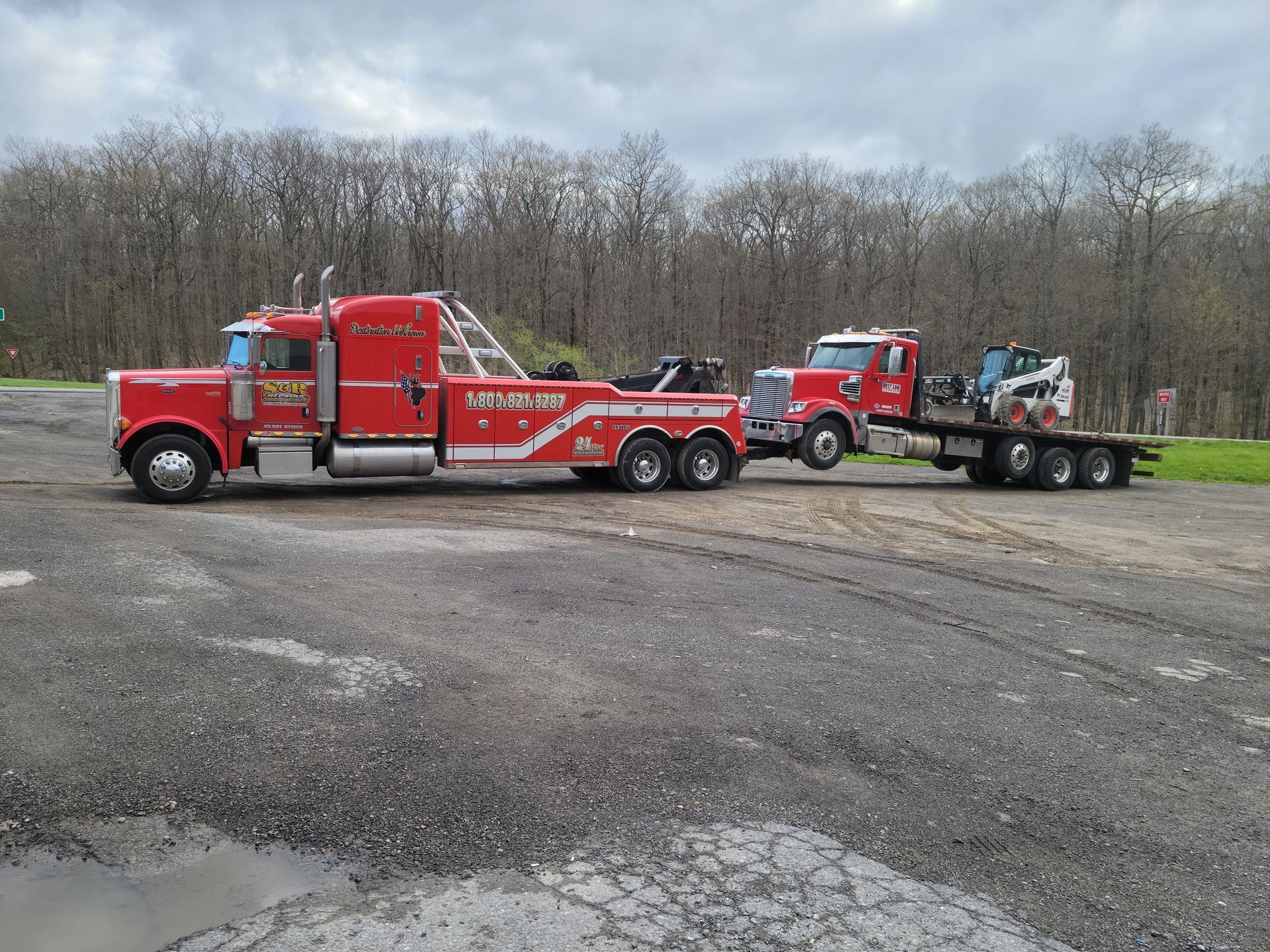 Red tow trucks and flatbed trailer parked on gravel in a wooded lot under cloudy skies