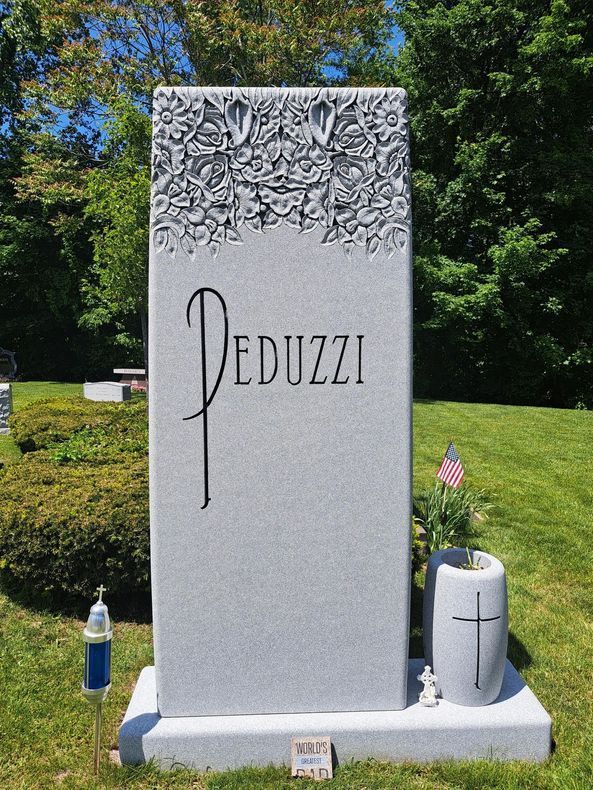 Decorated gravestone with the name Peduzzi in a grassy cemetery, with flowers and a small U.S. flag.