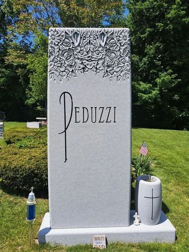 Gray memorial headstone engraved “Peduzzi” in a green cemetery, with flowers carved at the top.