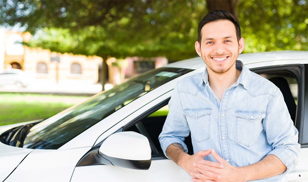 A smiling man in a light blue denim shirt leans against the side of a white car in a sunlit outdoor setting A smiling man in a light blue denim shirt leans against the side of a white car in a sunlit outdoor setting