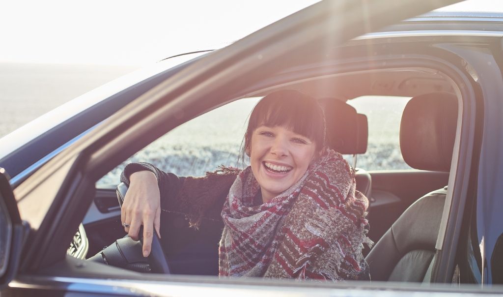 Learn More About Automotive Insurance A young woman with dark bangs and a plaid scarf smiles brightly while sitting in the driver's seat of a car, backlit by strong sunlight streaming through the window
