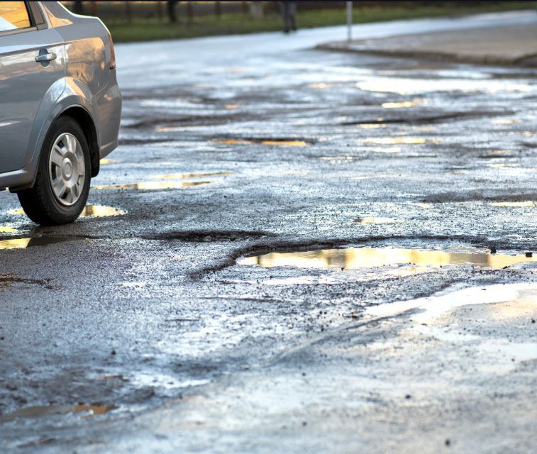 A car driving on a severely damaged asphalt road with multiple large, water-filled potholes