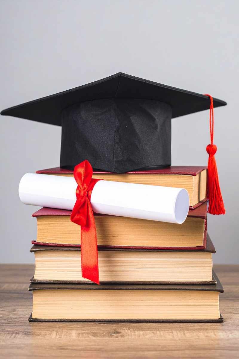 A black graduation cap with a red tassel sits on top of a stack of four hardcover books, with a roll
