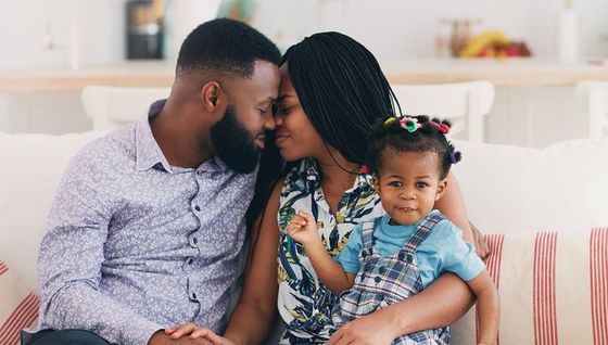 A Black man and woman sit on a sofa, touching foreheads affectionately while holding their young daughter who smiles at the camera