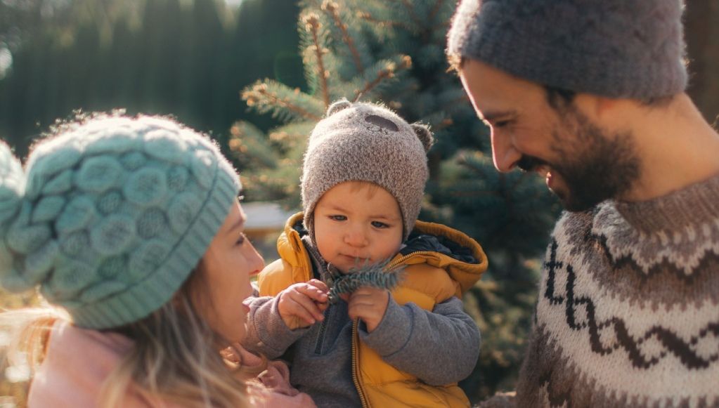 Learn More About Home Insurance A mother and father lovingly gaze at their toddler, who is wearing a bear-eared hat and holding a pine sprig