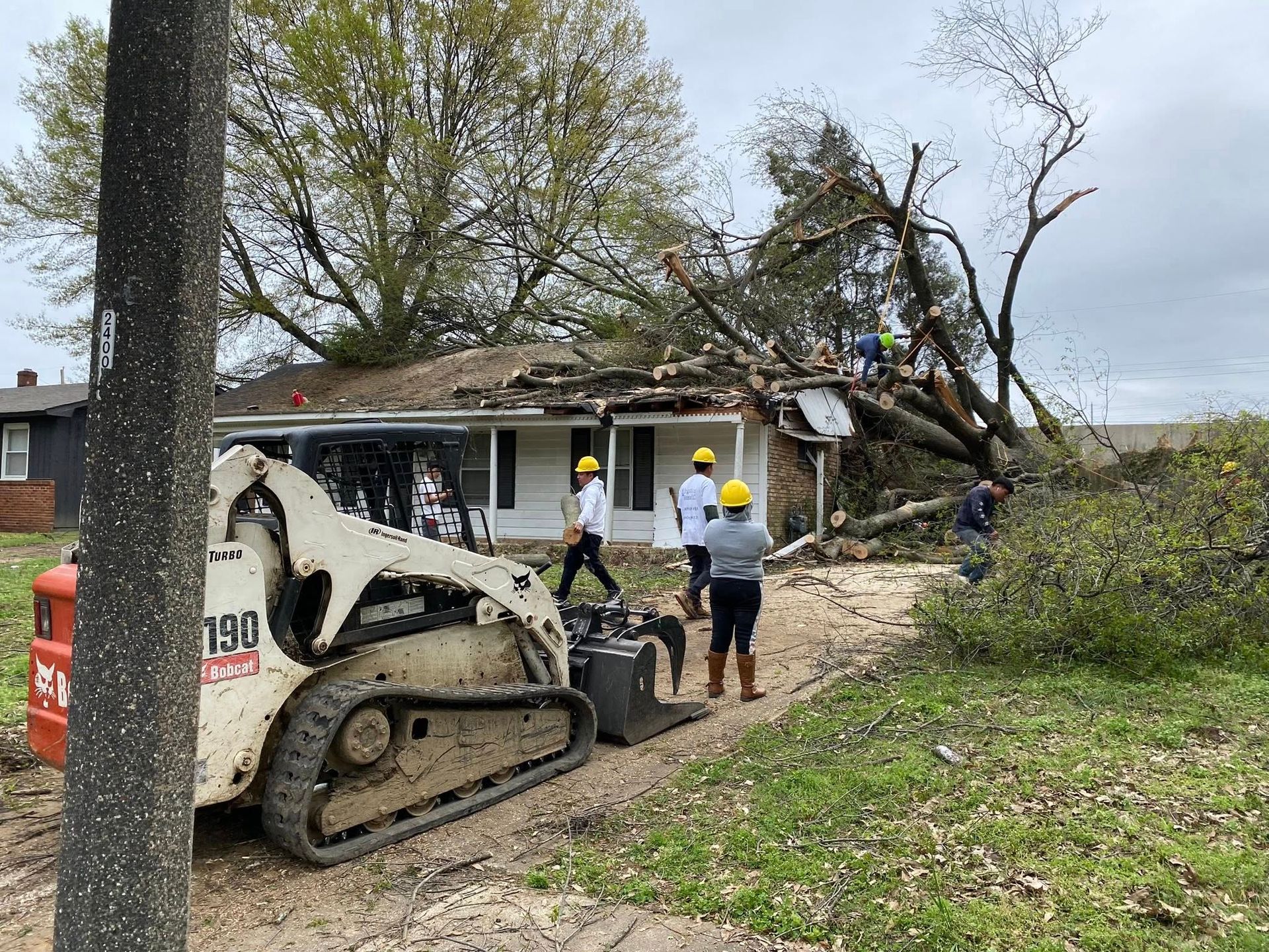 Workers use a skid steer to clear a large tree that has fallen onto the roof of a house after a storm.