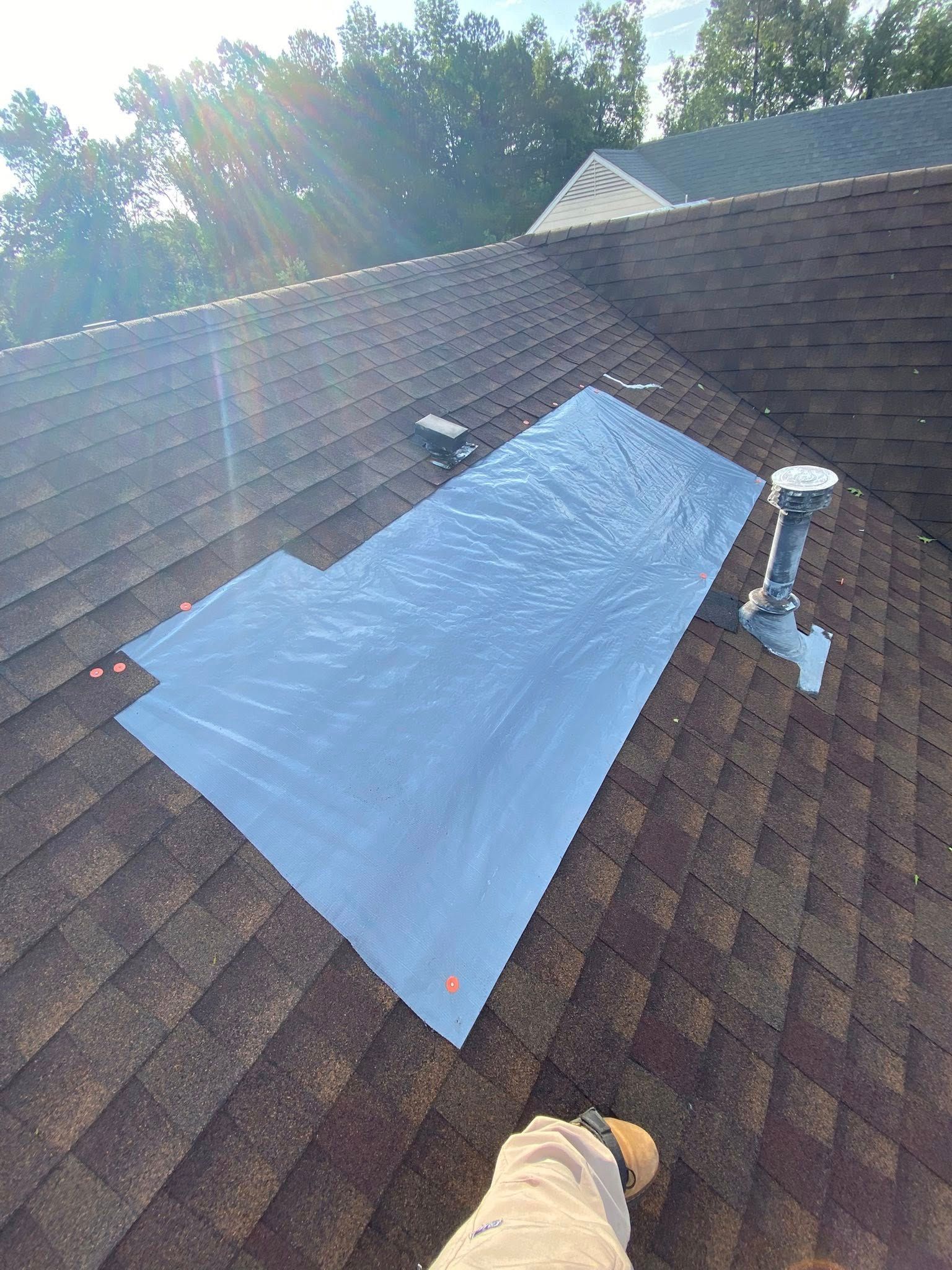 A blue waterproof tarp is patched over a section of brown asphalt shingles on a residential roof near a plumbing vent.