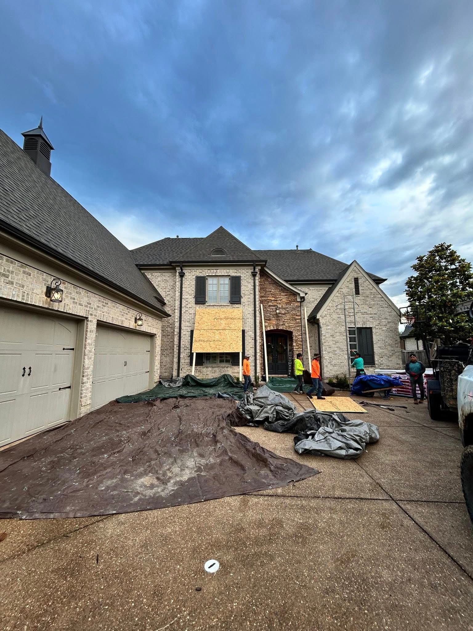 A construction crew works on the front exterior of a large, two-story brick home with a plywood-covered window.