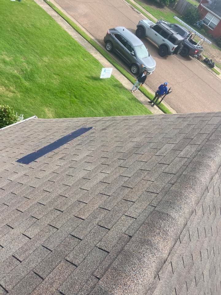A high-angle view of a shingled roof with two people standing near parked cars on the grass and pavement below.