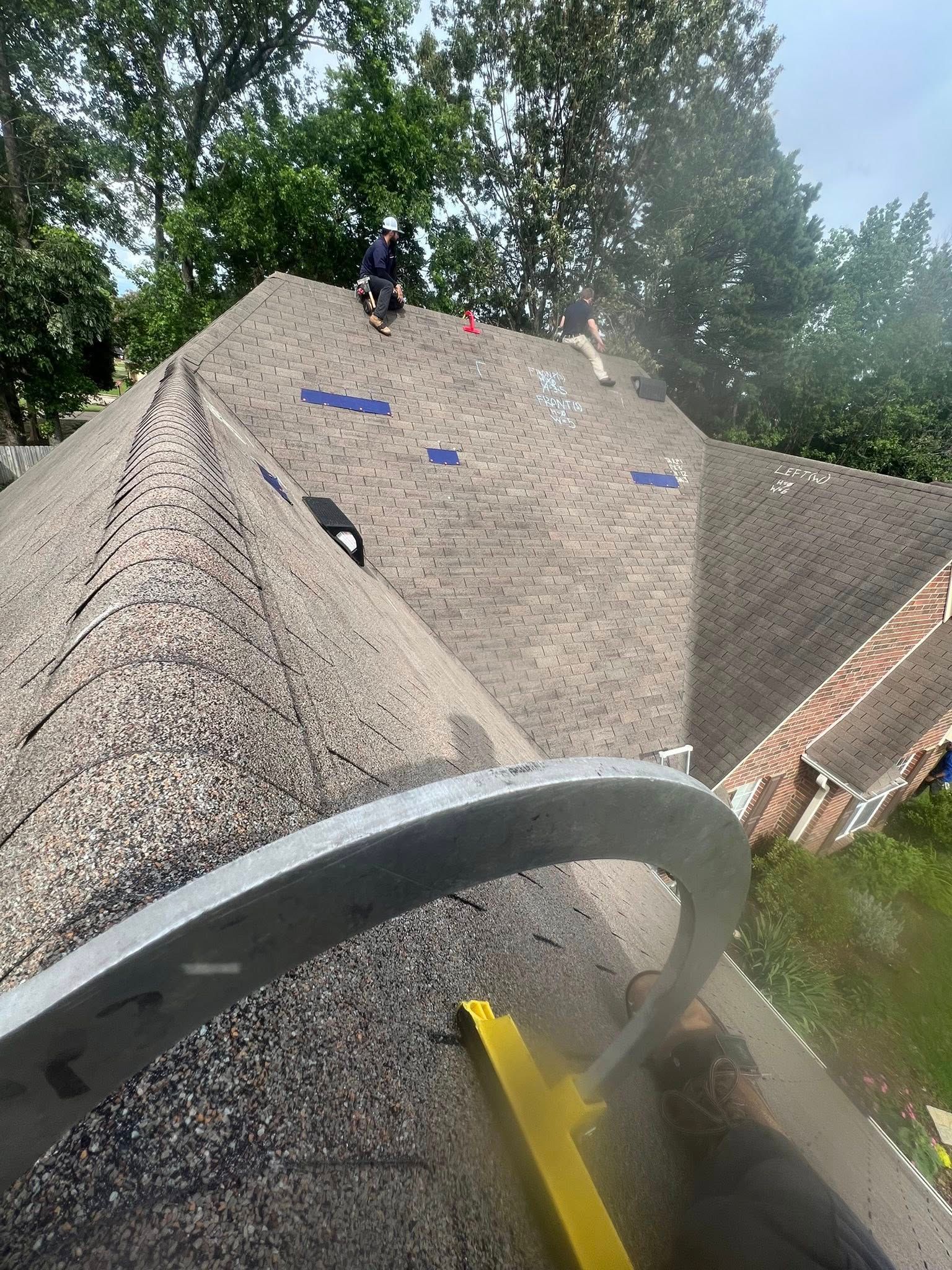A worker stands on a brown shingled roof while a curved yellow tool clears debris from the surface.