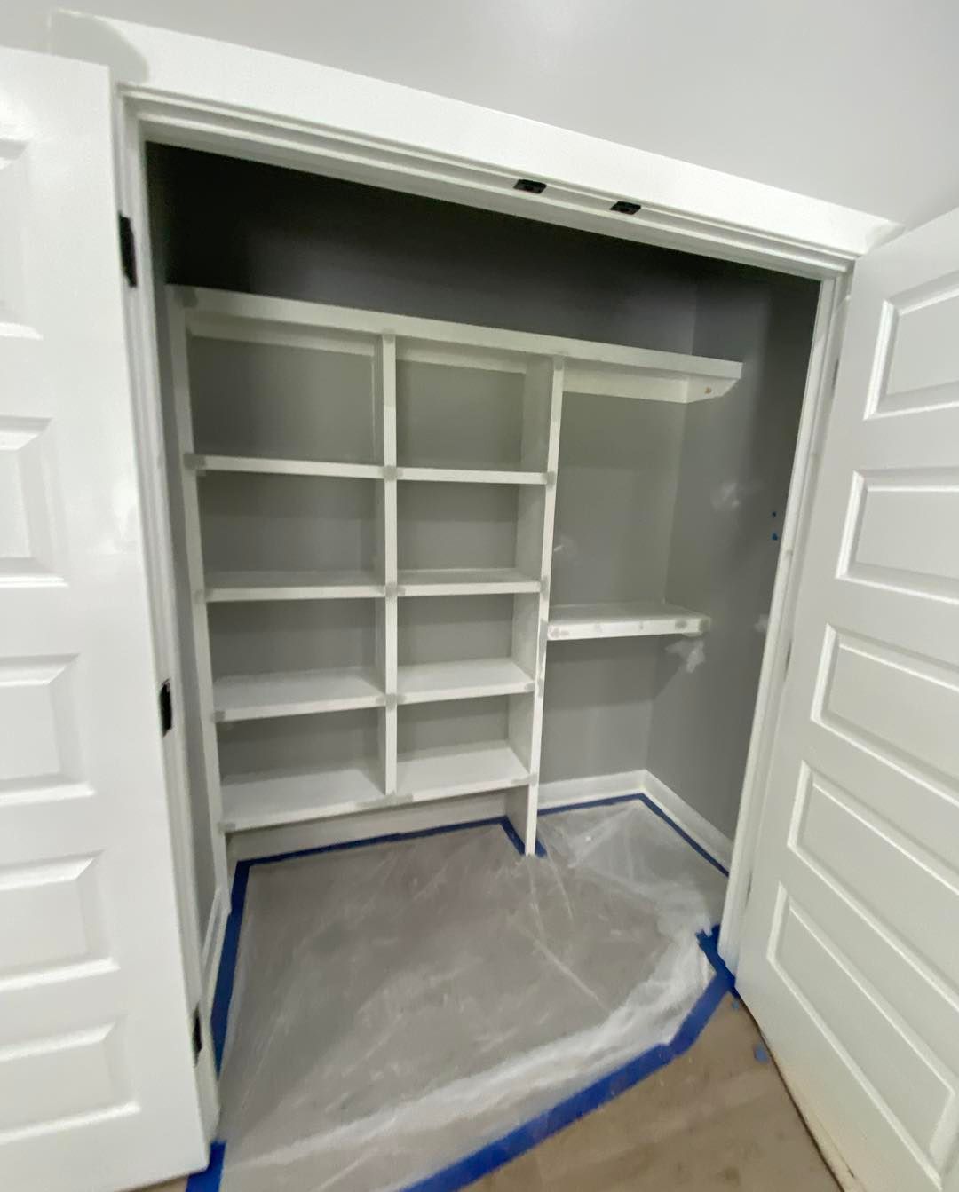 A white reach-in closet with built-in shelves and a side hanging rod, set against gray walls with plastic on the floor.