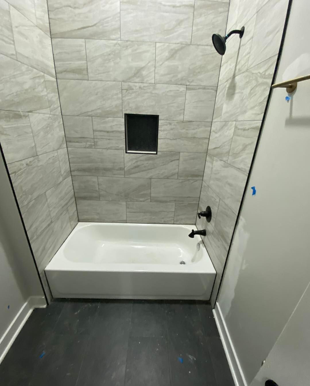 A bathroom featuring a white bathtub, gray tiled walls, a black showerhead, faucet, and a small recessed wall niche.