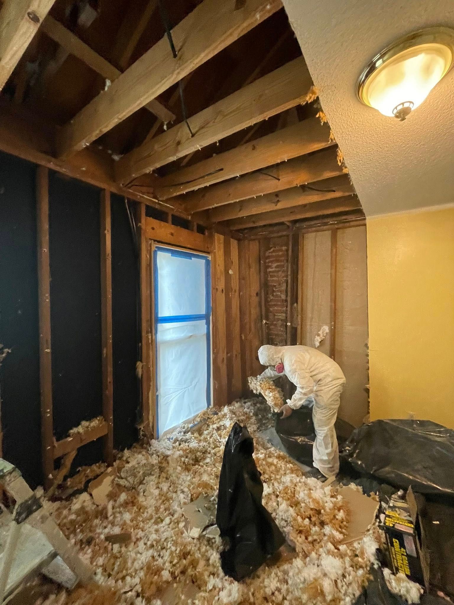 A person in a protective suit works in a room with exposed wall studs, ceiling joists, and scattered insulation debris.