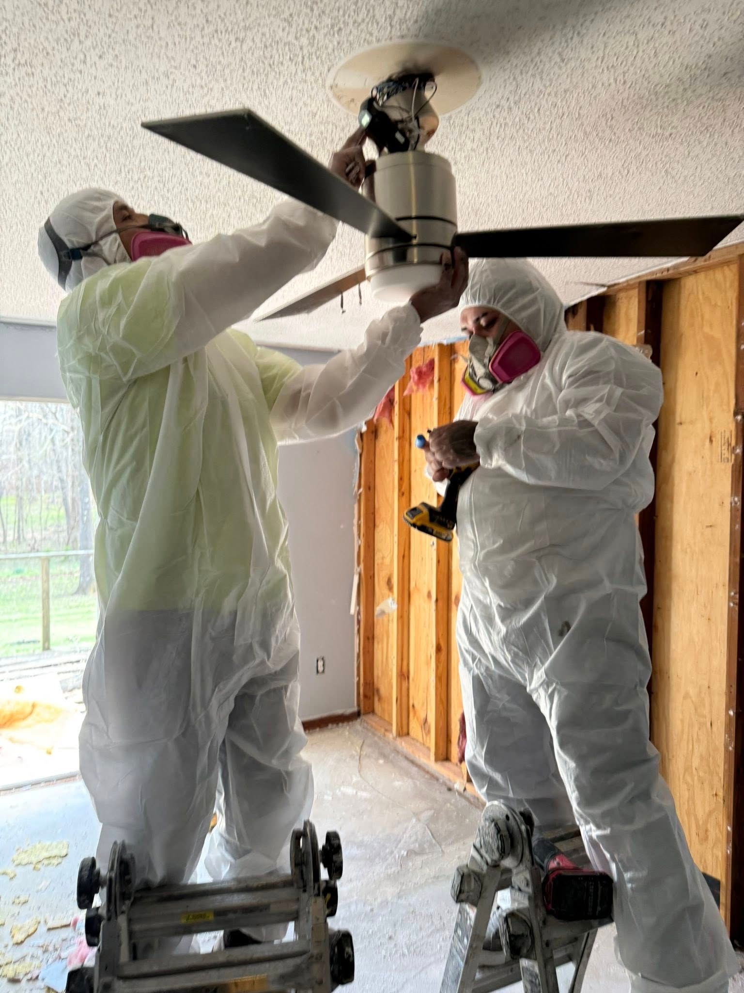 Two people in protective suits and respirators use ladders to remove a ceiling fan from a room under construction.
