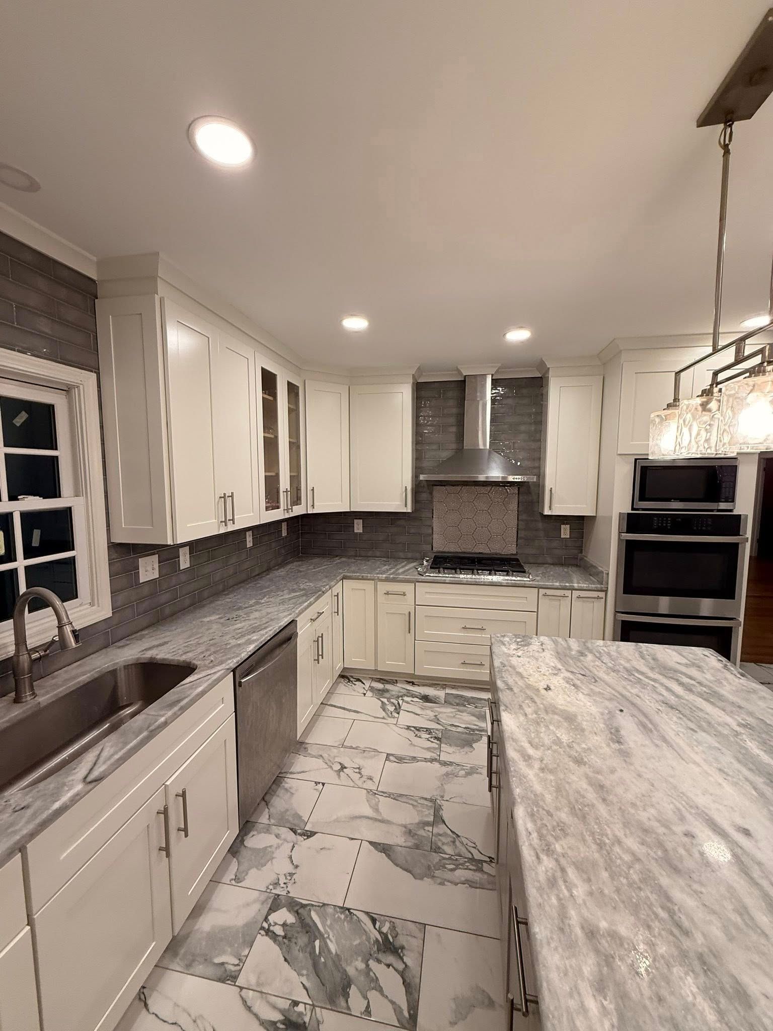 A bright, modern kitchen with white cabinets, gray granite countertops, stainless steel appliances, and patterned flooring.