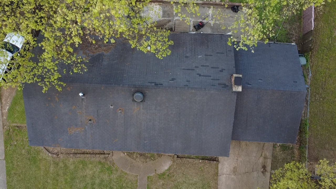 A top-down aerial view of a rectangular house roof with dark shingles surrounded by trees and a green lawn.