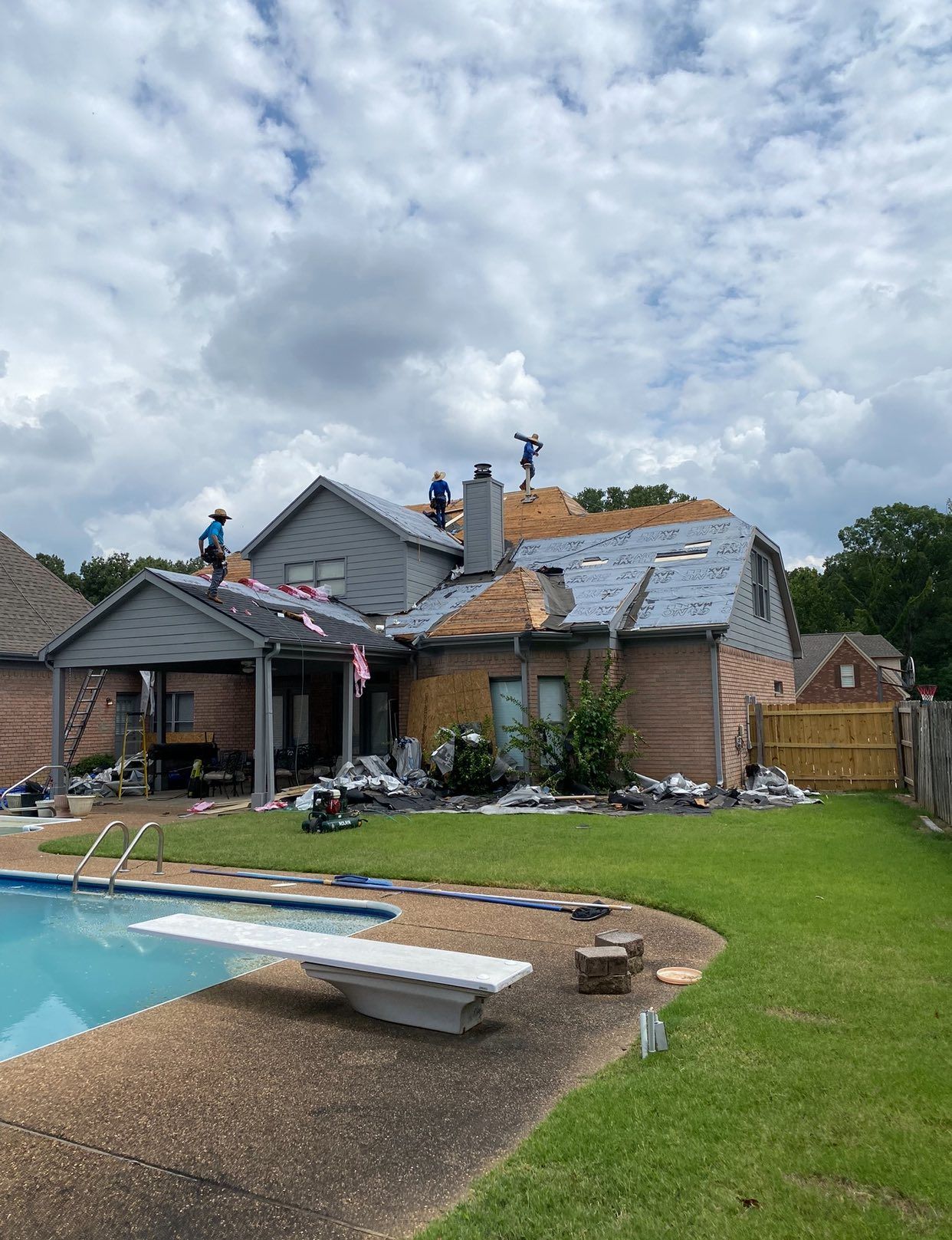 Workers remove old roofing from a residential house with a backyard swimming pool under a cloudy sky.