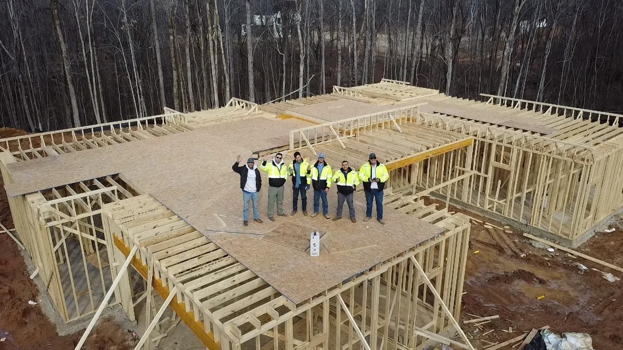 Six people in high-visibility vests stand on the wooden frame of a house under construction near a forest.