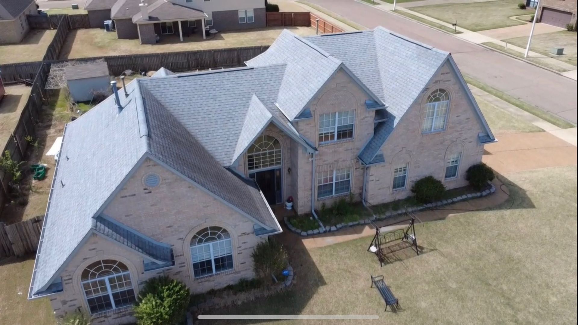 An aerial view of a two-story brick suburban house with a gray shingled roof, set in a neighborhood with a fenced yard.